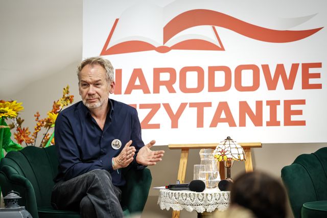Na zdjęciu znajduje się siedzący na fotelu znajdującym się na scenie, po prawej stronie stoi stolik z lampką, a za mężczyzną biała tablica z czerwonym napisem Narodowe Czytanie.