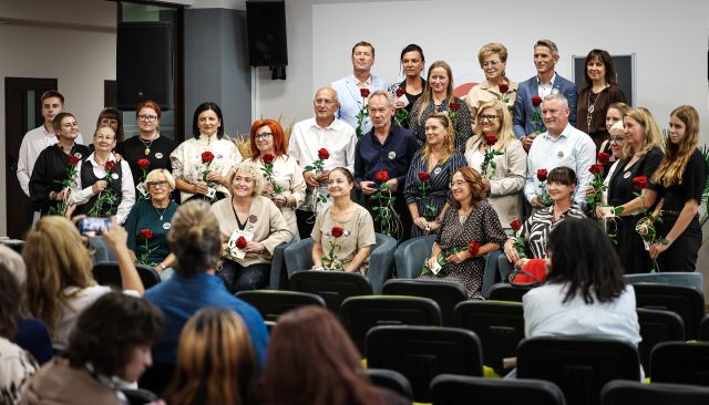 Na zdjęciu znajduje się około 25 osób stojących do zdjęcia, na scenie i przed sceną z różami, przed nimi znajdują się krzesła oraz widownia.