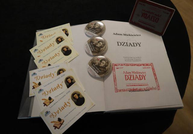 Na zdjęciu na stoliku znajduje się otwarta książka Dziadów Adama Mickiewicz wraz z pieczęcią Narodowego Czytania, obok leża trzy przypinki z Mickiewiczem, oraz małe podziękowania z Narodowego Czytania wraz z Adamem Mickiewiczem. W prawym górnym rogu książki znajduje się pieczęć.