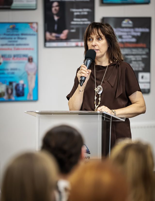 Na zdjęciu znajduje się kobieta z mikrofonem stojąca przy mównicy. W tle na ścianach znajdują się plakaty ze spotkań.