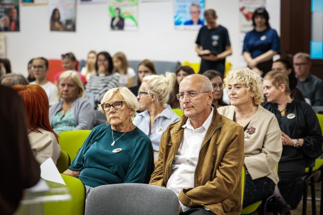 Na zdjęciu znajdują się zaproszeni goście na holu biblioteki, w centrum znajduje się kobieta wraz z mężczyzną. W tle znajdują się dwie stojące osoby oraz plakaty na ścianach.