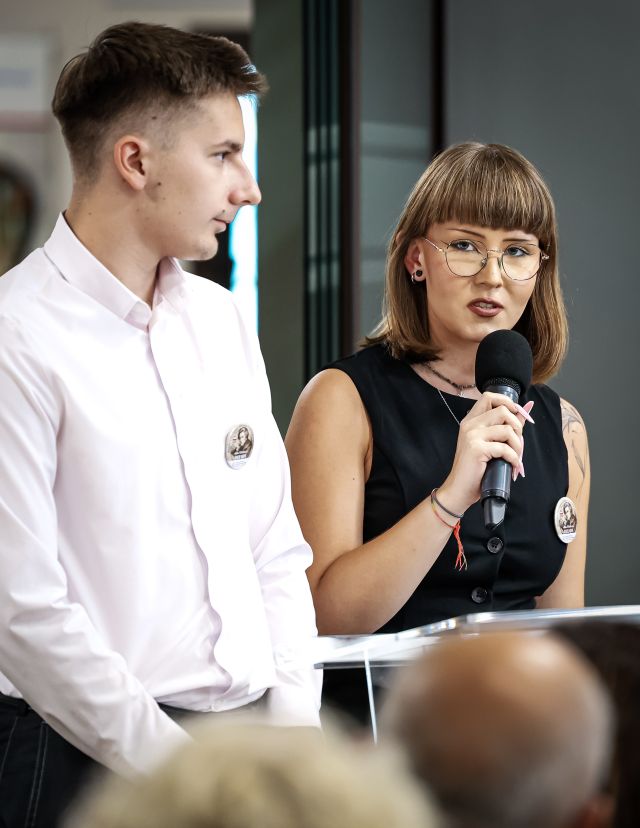 Na zdjęciu znajduje się chłopak oraz dziewczyna z mikrofonem przy mównicy. W tle znajduje się przesuwana szklana ścianka.