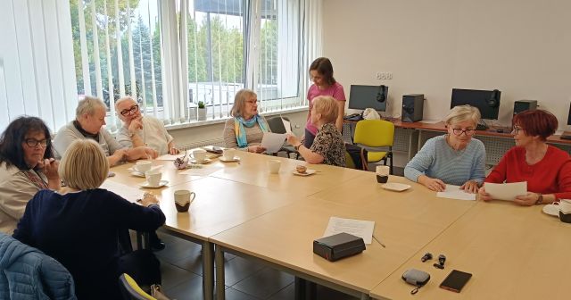 na zdjęciu seniorki rozmawiające ze sobą w parach, przy dwóch stoi prowadząca warsztaty, w tle komputery