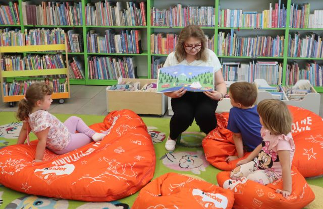 Na zdjęciu znajduje się kobieta pokazująca siedzącym dzieciom na pufie przed nią ilustracje z książki. Dzieci siedzą na pomarańczowych pufach na dywanie, a w tle znajdują się półki z książkami.
