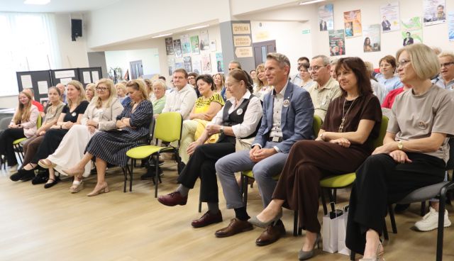 Na zdjęciu znajduje się widownia na holu biblioteki, około 100 osób. W tle znajdują się plakaty na ścianach.