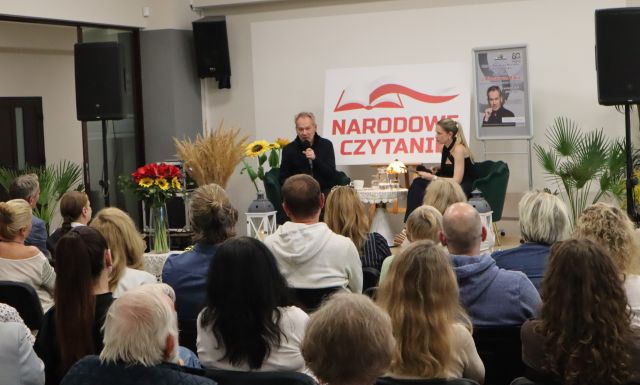 Na zdjęciu znajduje się widownia na holu biblioteki, a przed nimi na scenie siedzi mężczyzna ora kobieta, za nimi znajdują się kwiaty, tablica z napisem Narodowe Czytanie oraz plakat promujący spotkanie z Mirosławem Baką.