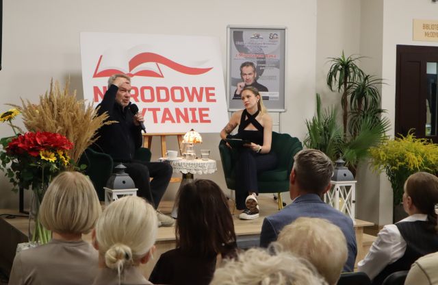 Na zdjęciu na scenie przed widownią siedzi mężczyzna oraz kobieta, pomiędzy nimi stoi stolik z lampką oraz wodą, za nimi znajduje się tablica z Narodowego Czytania oraz plakat promujący spotkanie z Mirosławem Baką. Na scenie znajdują się również kwiaty.