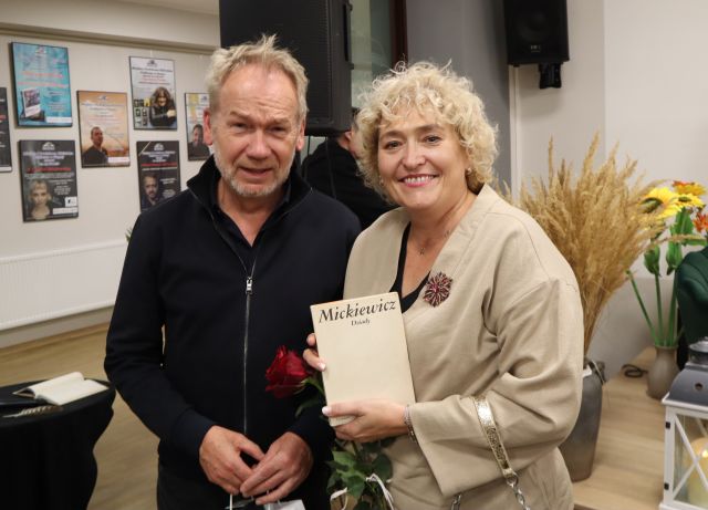 Na zdjęciu znajduje się kobieta stojąca do zdjęcia z mężczyzną, w ręce tryma książkę Mickiewicza. W tle znajdują się kwiaty oraz plakaty na ścianie.
