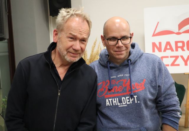 Na zdjęciu znajduje się dwóch mężczyzn stojących do wspólnego zdjęcia, w tle znajduje się biała tablica z napisem Narodowe Czytanie w czerwonym kolorze.