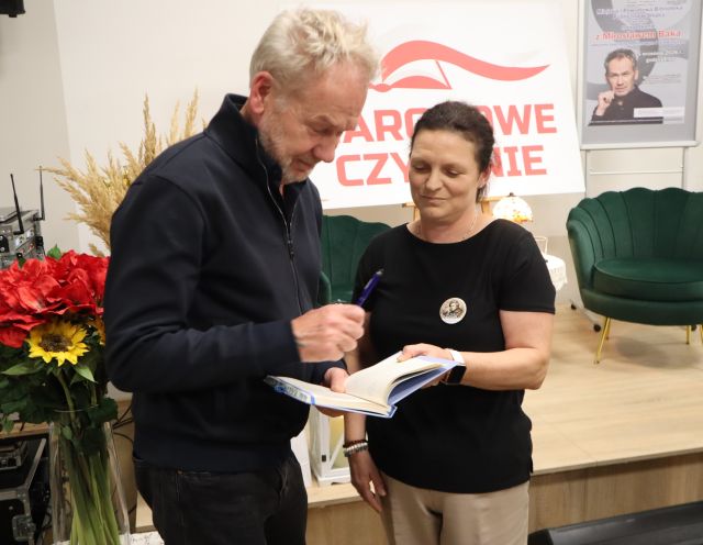 Na zdjęciu znajduje się kobieta wraz z mężczyzną, który podpisuje się w zeszycie, za nimi znajdują się kwiaty oraz scena z fotelami, białą tablicą z napisem Narodowe Czytanie oraz plakatem promującym spotkanie z Mirosławem Baką.