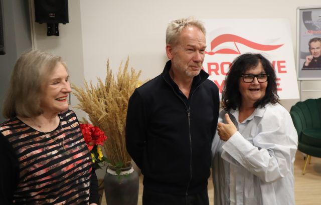 Na zdjęciu znajdują się dwie kobiety stojące do zdjęcia z mężczyzną. W tle znajdują się kwiaty oraz biała tablica z czerwonym napisem Narodowe Czytanie i plakatem promującym spotkanie z Mirosławem Baką.