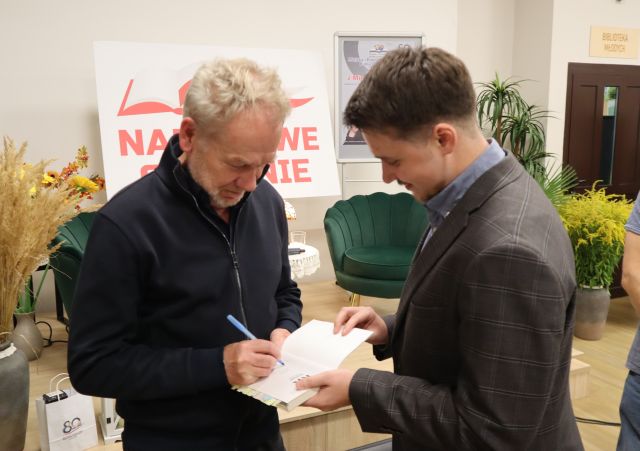Na zdjęciu znajduje się dwóch mężczyzn, jeden podpisuje się drugiemu w zeszycie. W tle znajdują się fotele, kwiaty, biała tablica z napisem oraz drzwi obok sceny.