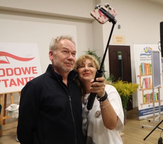 Na zdjęciu znajduje się mężczyzna oraz kobieta, która robi im wspólne zdjęcia za pomocą selfie stick'a. W tle znajduje się scena z białą tablicą z czerwonym napisem, drzwi oraz baner z książkami i logiem biblioteki.