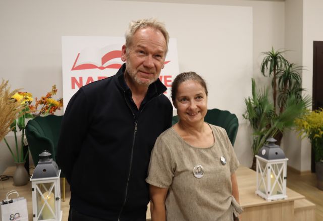 Na zdjęciu znajduje się kobieta stojąca do zdjęcia z mężczyzną. W tle znajduje się scena z fotelami, kwiatami, dwoma lampionami oraz białą tablicą z czerwonym napisem.