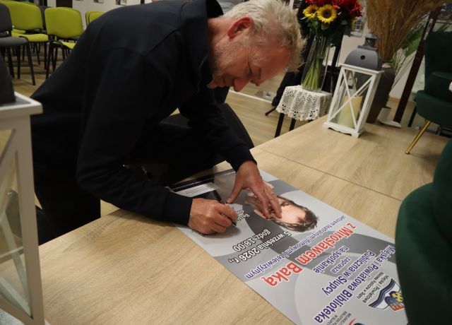 Na zdjęciu znajduje się mężczyzna, który się pochyla i podpisuje plakat promujący spotkanie. W tle znajduje się lampion, kwiaty na stoliku.