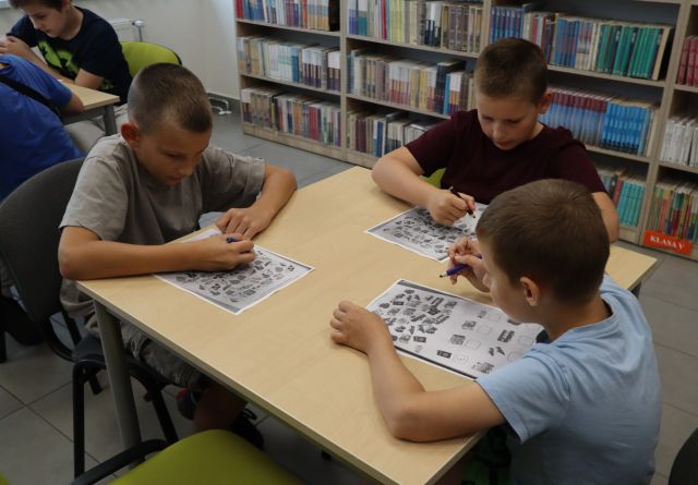 Na zdjęciu znajduje się trzech chłopców przy stoliku i rozwiązują karty pracy, w tle znajdują się półki z książkami.