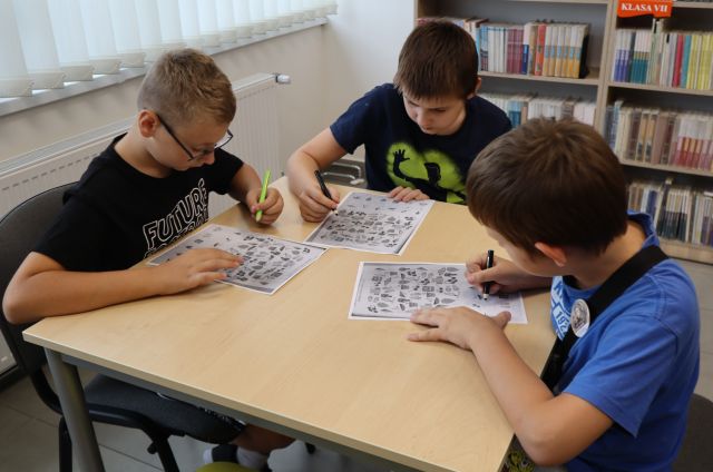 Na zdjęciu znajduje się trójka chłopców, którzy rozwiązują karty pracy przy stoliku. W tle znajdują się półki z książkami oraz okna.