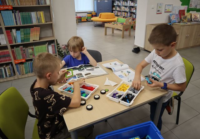 Na zdjęciu znajduje się trójka chłopców przy stoliku, dwóch z nich układa klocki LEGO, a jeden czyta książkę. W tle znajdują się półki z książkami.