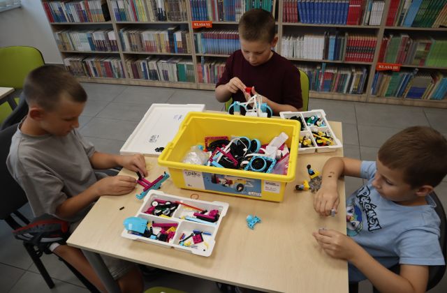 Na zdjęciu znajduje się trójka chłopców przy stoliku, którzy wspólnie układają klocki, które znajdują się w plastikowym pojemniku na stoliku. W tle znajdują się półki z książkami.