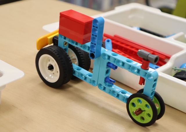 Na zdjęciu znajduje się zbudowany z klocków LEGO rower stojący na stoliku, za nim znajduje się pudełko z klockami.