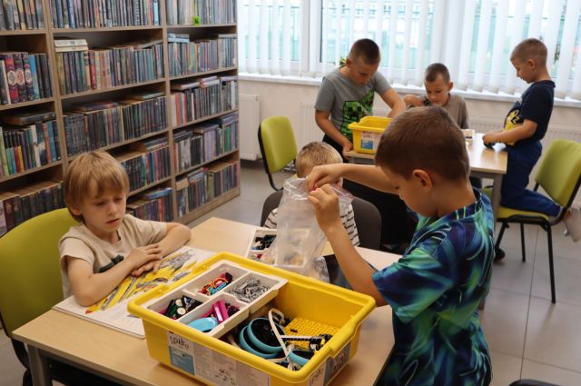 Na zdjęciu znajduje się sześciu chłopców przy dwóch stolikach po trzy osoby i układają z klocków LEGO znajdujących się na stole chwytaki. W tle znajdują się półki z książkami i okna.