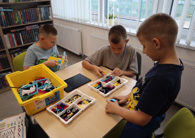 Na zdjęciu znajduje się 3 chłopców przy stoliku i wspólnie układają klocki LEGO według znajdującej się na tablecie instrukcji. W tle znajdują się półki z książkami oraz okna.