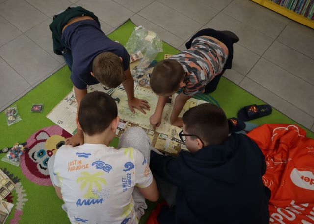Na zdjęciu znajduje się czwórka chłopców, którzy grają w grę planszową na dywanie, obok nich leży pufa.
