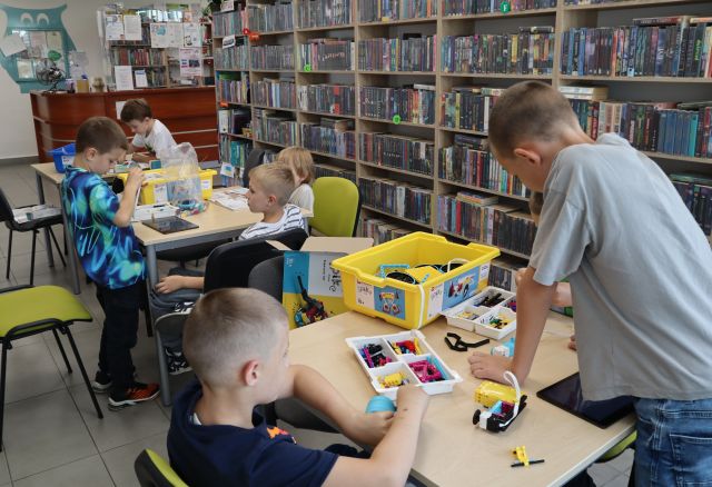 Na zdjęciu znajduje się 7 chłopców przy trzech stolikach, przy których układają klocki LEGO. W tle znajdują się półki z książkami oraz biurko biblioteczne.