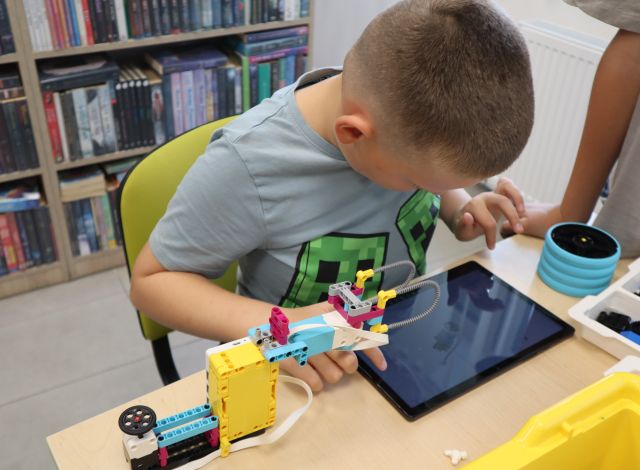 Na zdjęciu znajduje się chłopiec przy stoliku, przed nim leży tablet z instrukcją oraz chwytak wykonany z klocków LEGO. W tle znajdują się półki z książkami.