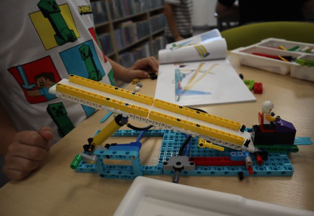 Na zdjęciu znajduje się stojąca na stoliku, wykonana z klocków LEGO skocznia narciarska, w tle znajduje się chłopiec, reszta klocków, instrukcja oraz półki z książkami.