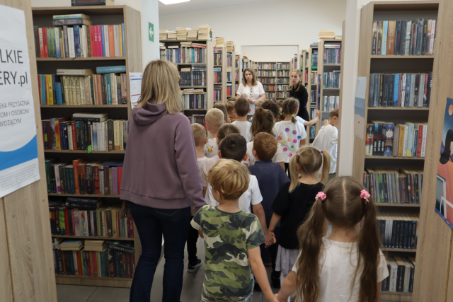 Na zdjęciu znajduje się grupa przedszkolaków z trzema paniami w sali bibliotecznej między półkami z książkami.