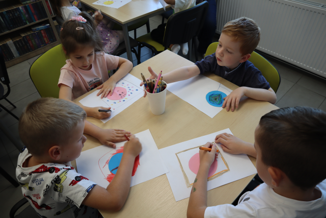 Na zdjęciu znajduje się czwórka dzieci przy stoliku i rysują obrazki z wykorzystaniem kropki, na środku stolika stoją kredki.
