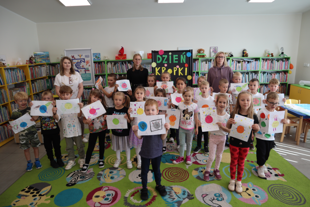 Na zdjęciu znajduje się 23 dzieci, które stoją do zdjęcia ze swoimi pracami z kropek, za nimi stoją dwie kobiety. W tle znajdują się półki z książkami oraz czarna tablica z napisem Dzień Kropki.