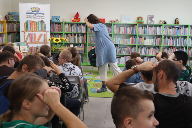 Na zdjęciu znajdują się dzieci i młodzież na krzesłach, przed nimi stoi kobieta bokiem, ręce ma nad oczami tak jak widownia i lekko się nachyla w bok. W tle znajdują się półki z książkami, fotel, kwiaty oraz baner z logiem biblioteki i książkami.