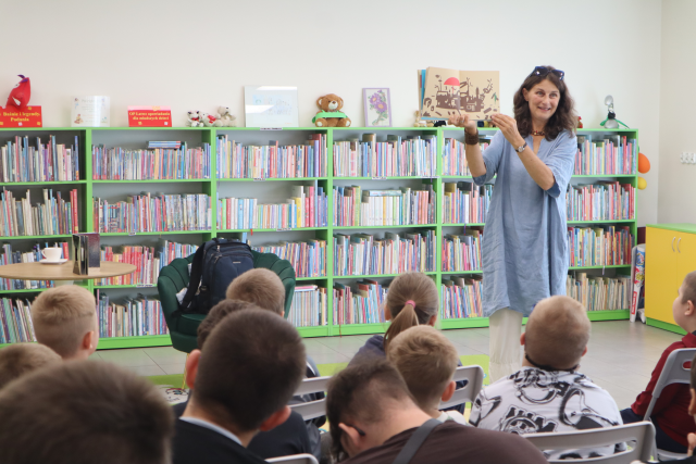 Na zdjęciu znajduje się kobieta w ręce trzyma książkę i pokazuje ją dzieciom, które siedzą przed nią. W tle znajduje się fotel, stolik z filiżanką oraz półki z książkami.