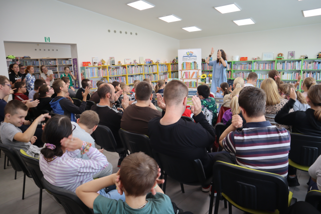 Na zdjęciu znajduje się około 50 dzieci i młodzieży, którzy siedzą na krzesłach i powtarzają ruchy za kobietą przed nimi, która klaszcze w dłonie. W tle znajdują się półki z książkami oraz baner z logiem biblioteki i książkami.