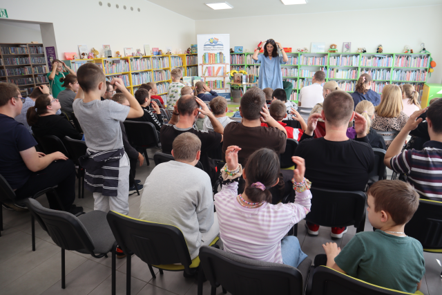 Na zdjęciu znajdują się dzieci i młodzież na widowni, ręce trzymają na poziomie głowie tak jak kobieta przed nimi. W tle znajdują się półki z książkami, stolik, baner z logiem biblioteki i książkami.