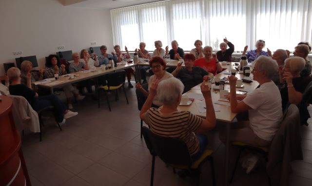 Na zdjęciu znajduje się około 25 kobiet przy złączonych stolikach w podkowę, ręce mają zgięte w łokciach i podniesione do góry. Na stolikach znajdują się kubki, termosy, ciasteczka. Warsztaty odbywają się w sali komputerowej, w tle znajdują się okna.