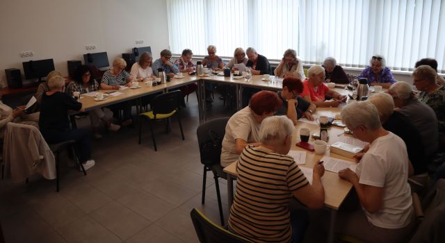 Na zdjęciu znajdują się kobiety przy złączonych stołach w podkowę i robią zadania, na stole znajdują się również kubki, termosy. Warsztaty odbywają się w sali komputerowej, w tle znajdują się okna.