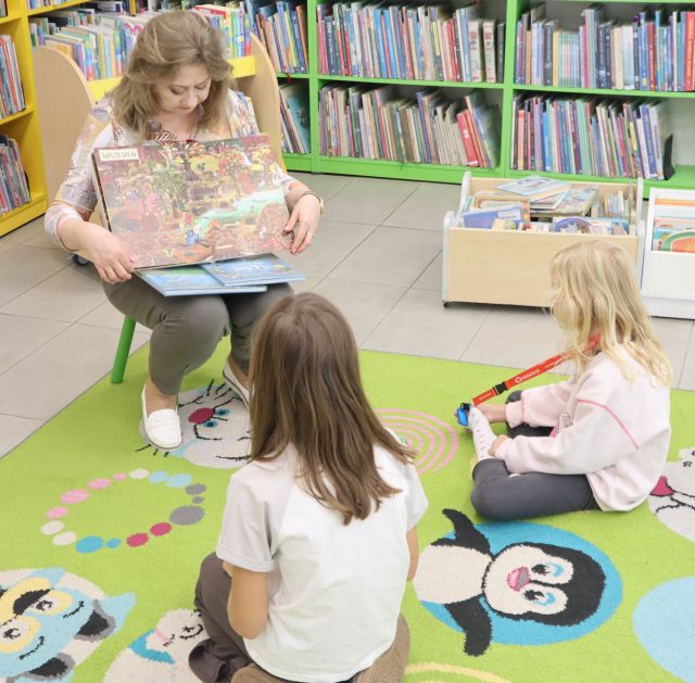 na zdjęciu na krzesełku siedzi bibliotekarka z książką, przed nią na kolorowym dywanie dwójka dzieci, w tle regały z książkami