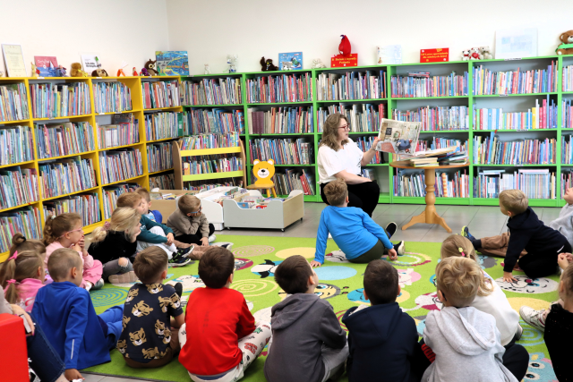 na zdjęciu bibliotekarka z książką siedzi na krzesełku przed przedszkolakami siedzącymi na kolorowym dywanie, w tle regały z książkami
