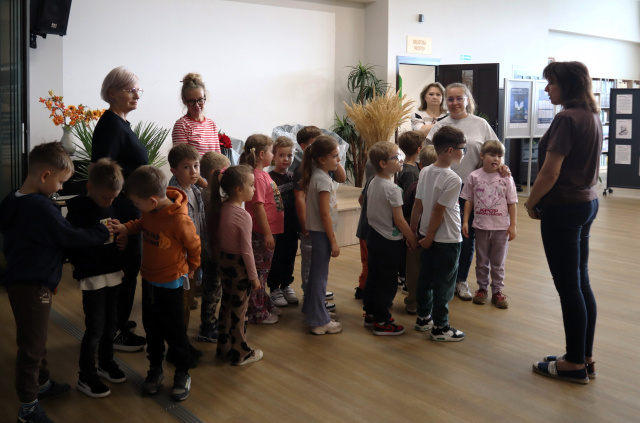 na zdjęciu dyrektorka biblioteki witająca w holu biblioteki grupę przedszkolaków, w tle drzwi i plakaty na ścianach