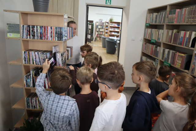 na zdjęciu przedskolaki i kobieta stoją przed regałem obrotowym z audiobookami