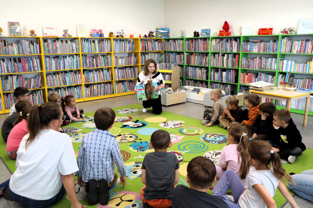 na zdjęciu bibliotekarka z książką klęczy na kolorowym dywanie, przed nią w okręgu siedzą na dywanie przedszkolaki, w tle regały z książkami
