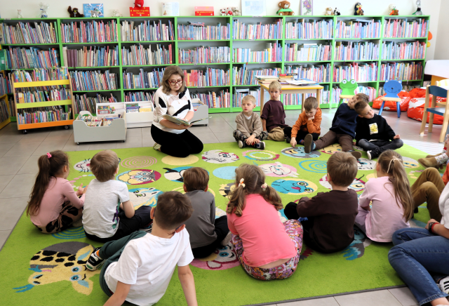 na zdjęciu bibliotekarka z książką klęczy na kolorowym dywanie, przed nią w okręgu siedzą na dywanie przedszkolaki, w tle regały z książkami