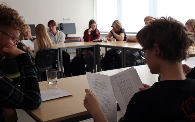 na zdjęciu młodzież siedząca przy stołąch ustawionych w literę u czyta z kartek