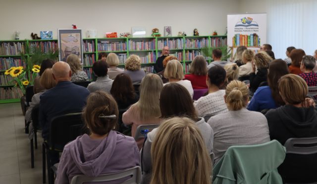 Na zdjęciu znajduje się widownia w sali bibliotecznej, przed nimi znajduje się kobieta i mężczyzna, plakat promujący spotkanie, baner z logiem biblioteki i książkami. W tle znajdują się półki z książkami oraz okna.