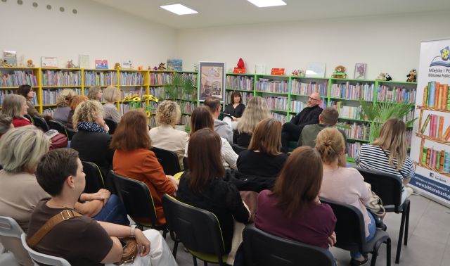 Na zdjęciu znajduje się widownia w sali bibliotecznej, przed nimi na fotelach siedzą mężczyzna i kobieta, po prawej stronie stoi baner z logiem biblioteki i książkami, po lewej stronie plakat promujący spotkanie. W tle znajdują się półki z książkami.