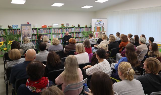 Na zdjęciu znajduje się widownia w sali bibliotecznej, przed nimi siedzi mężczyzna ora kobieta prowadząca spotkanie, po prawej stronie znajduje się baner z logiem biblioteki i książkami, a po lewej stronie plakat promujący spotkanie. W tle znajdują się półki z książkami oraz okna.