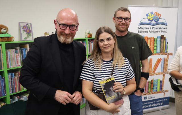 Na zdjęciu znajduje się dwóch mężczyzn i kobieta z książką i stoją do wspólnego zdjęcia. W tle znajdują się półki z książami i baner z logiem biblioteki i książkami.